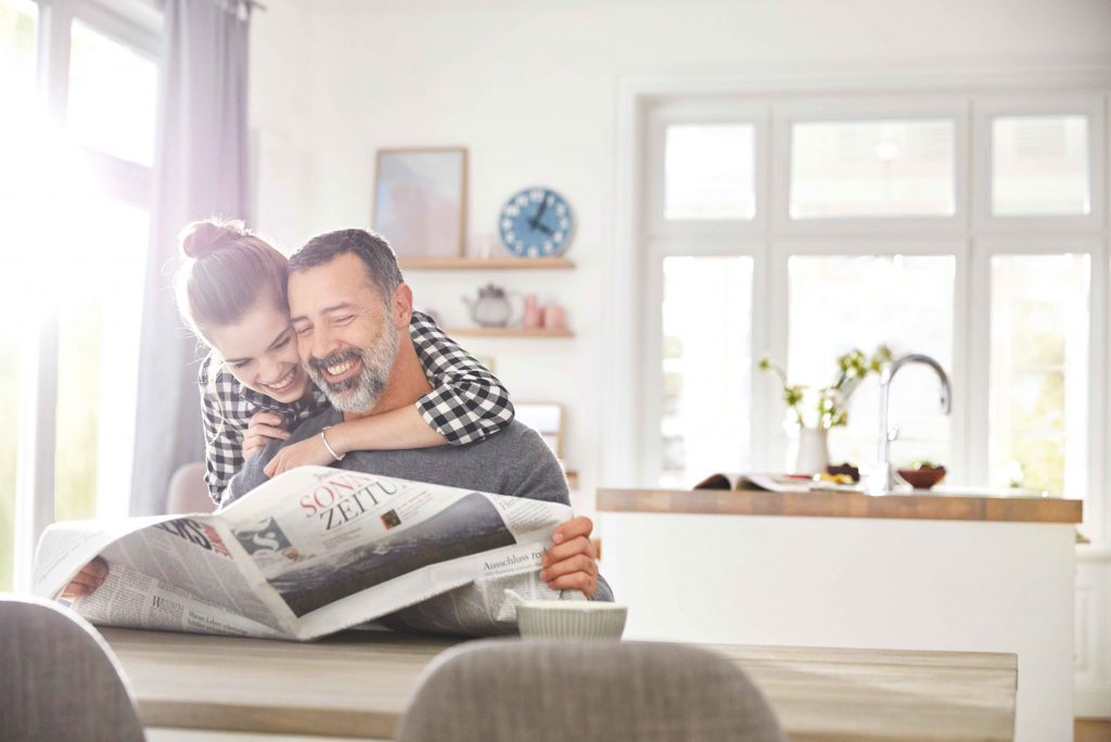 Eine Mädchen umarmt ihren Vater, der am Tisch sitzt und eine Zeitung liest